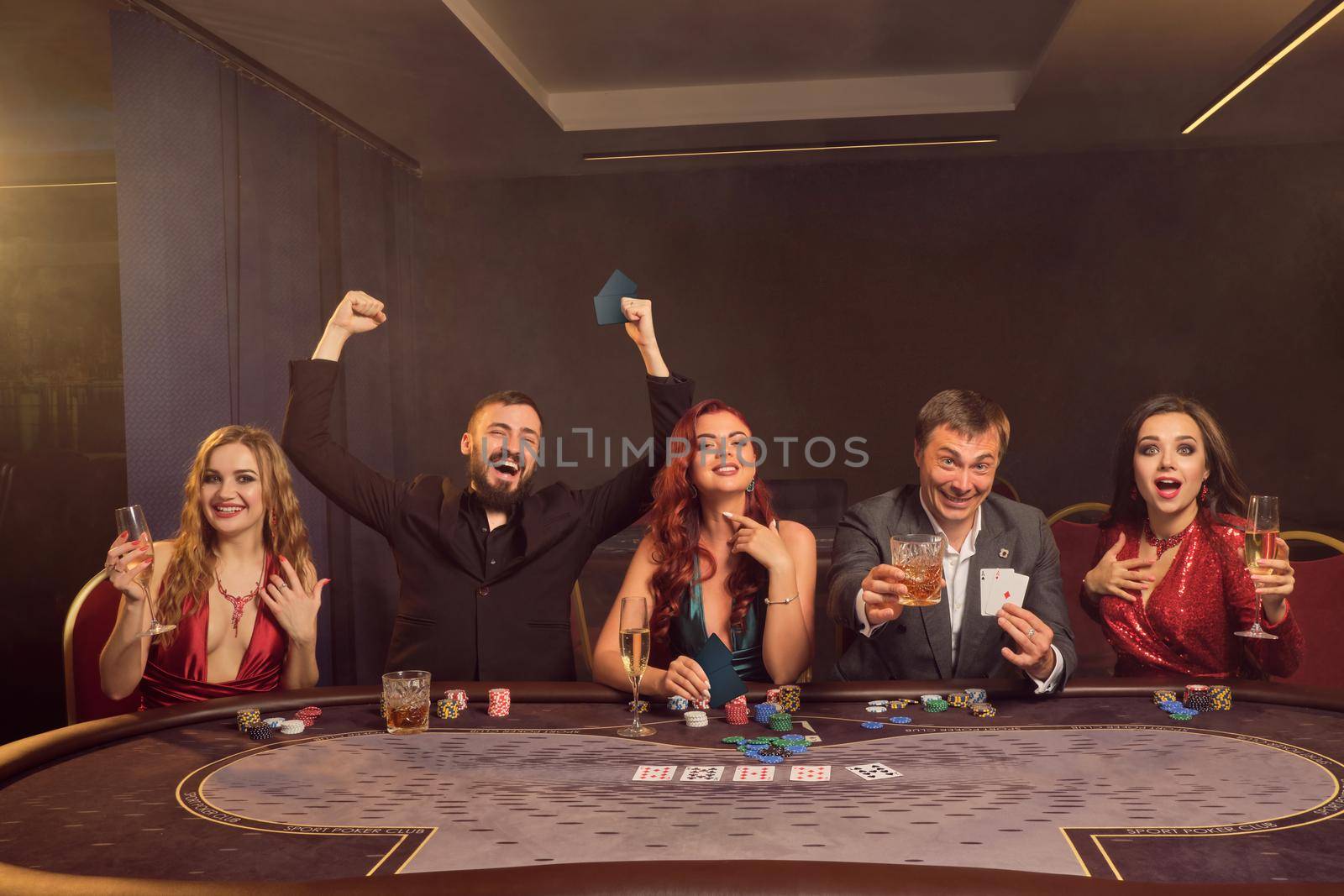 Optimistic classmates are playing poker at casino. They are celebrating their win, smiling and looking vey excited while posing at the table against a dark smoke background in a ray of a spotlight. Cards, chips, money, alcohol, gambling, entertainment concept.