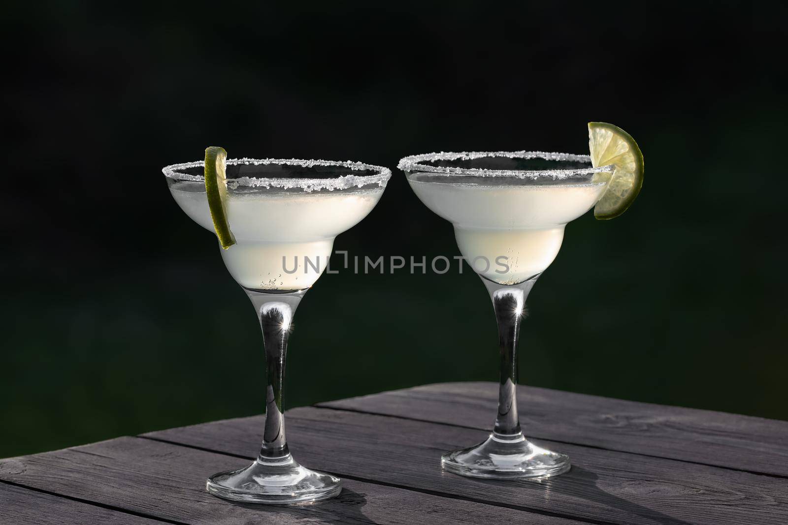 Two glasses with fresh margarita cocktail on a wooden table on a patio summer evening, selective focus by galsand