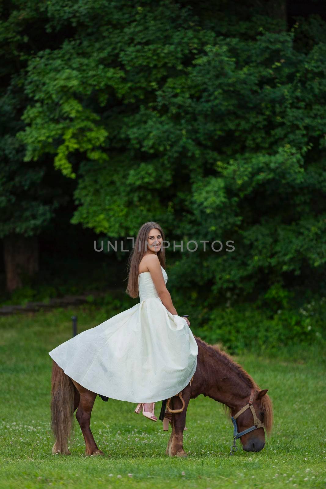 girl in a white dress rides a pony on a green lawn