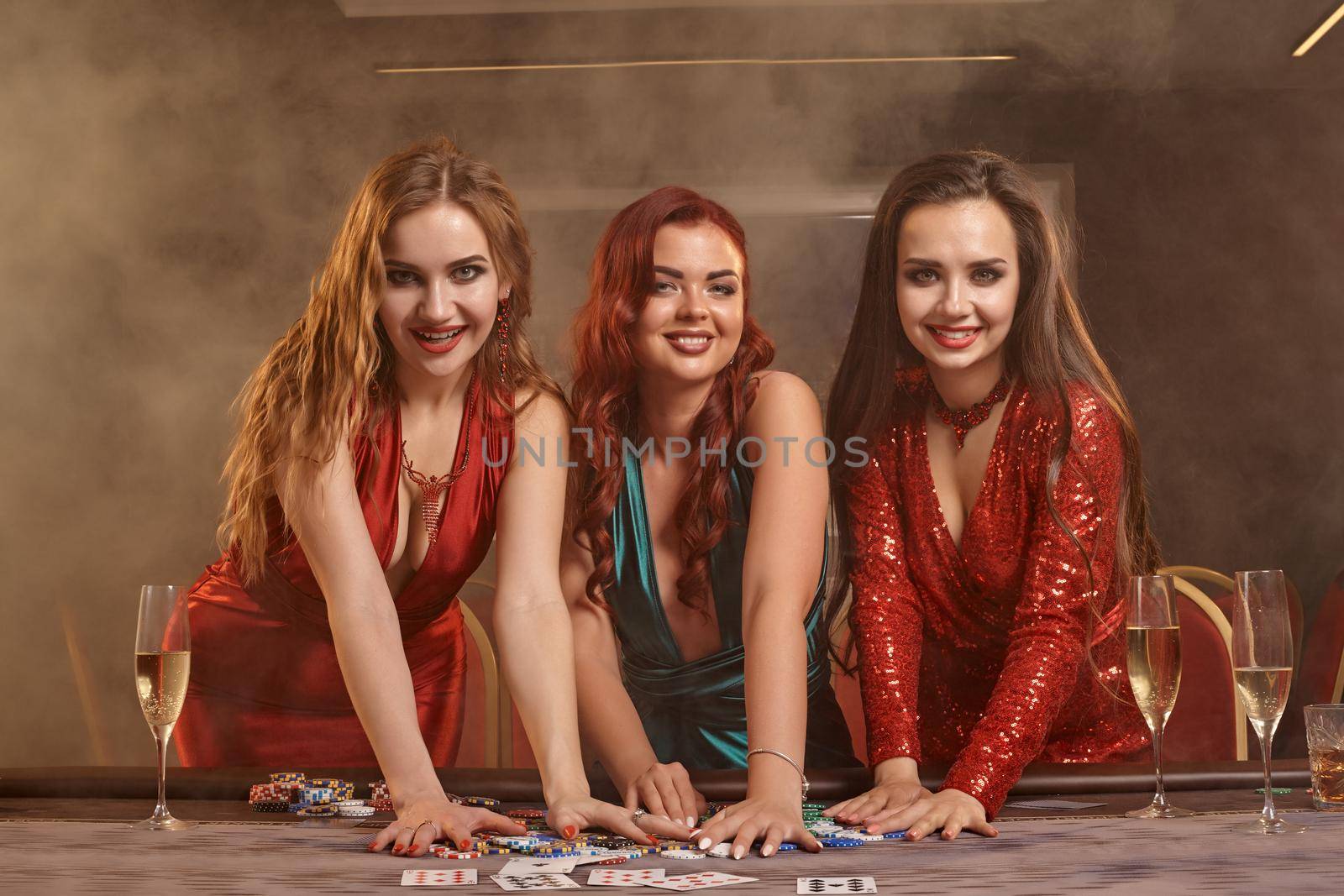 Three beautiful women are playing poker at casino. They are celebrating their win, looking at the camera and smiling, holding some chips and posing at the table against a smoke background. Cards, money, alcohol, gambling, entertainment concept.
