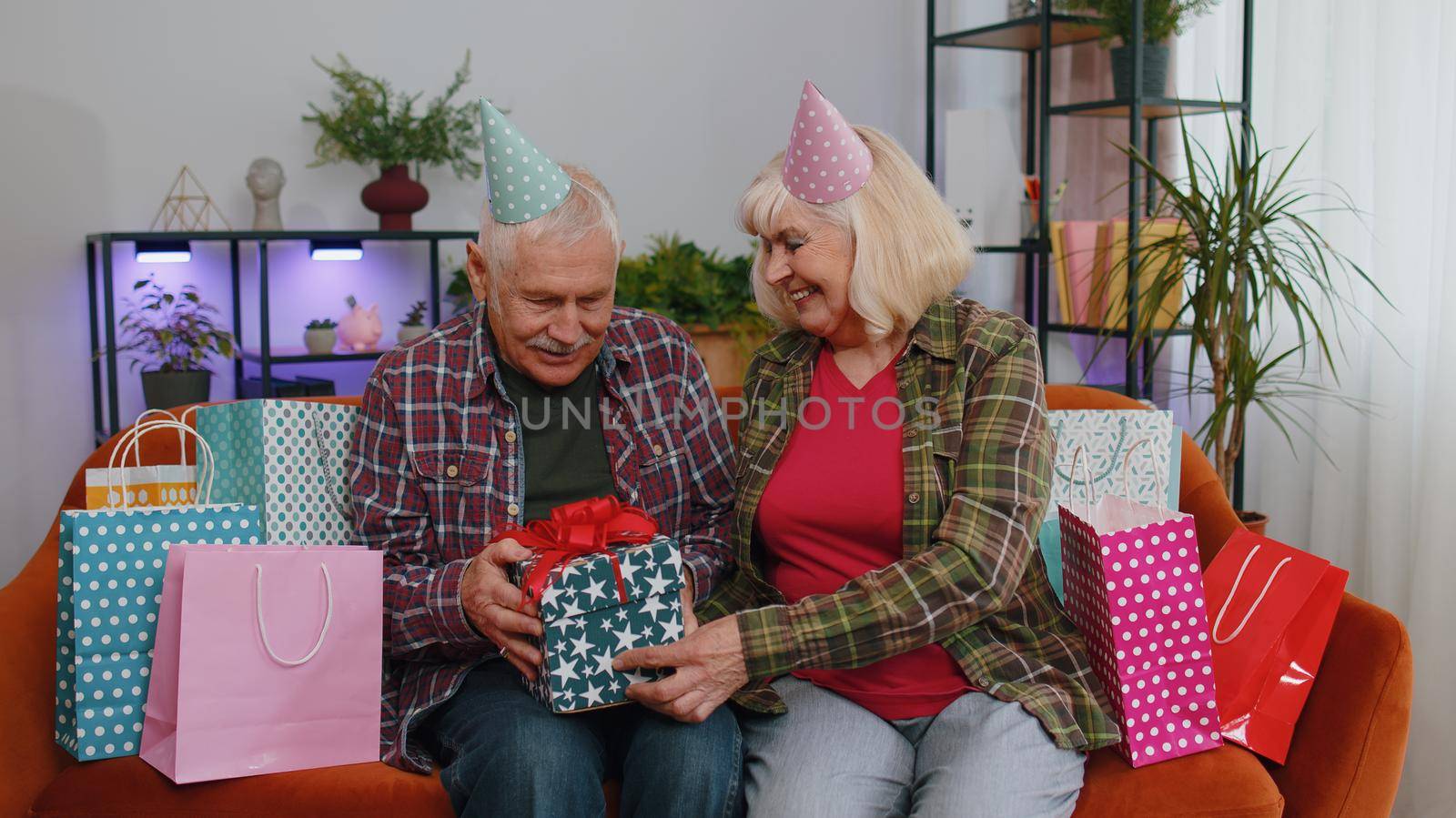 Happy old senior elderly family couple grandparents man woman celebrating birthday anniversary on couch at home. Mature grandmother wife giving gift box surprise present to her husband grandfather