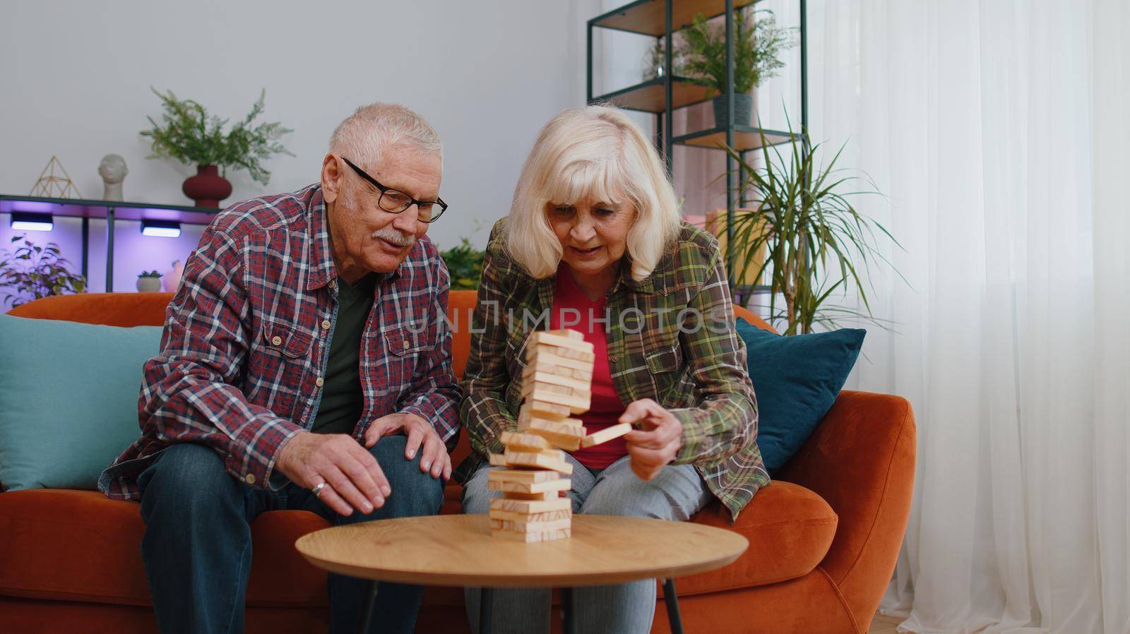 Old senior mature family couple grandparents man woman playing in blocks build game. Mature grandmother grandfather spending time together play board game hobby with wooden tower on couch at home