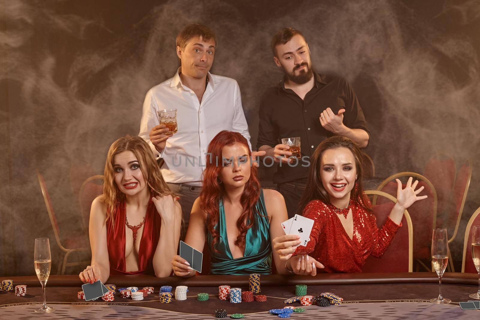 Agitated friends are playing poker at casino. They are celebrating their win, smiling and posing at the table against a dark smoke background. Cards, chips, money, alcohol, fortune, gambling, entertainment concept.