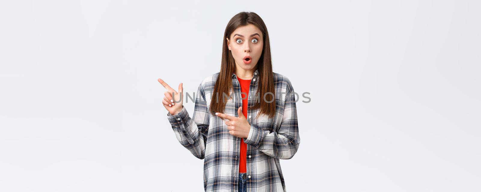 Lifestyle, different emotions, leisure activities concept. Impressed female customer, girl in casual checked shirt, say wow talking about news and pointing fingers left, white background.