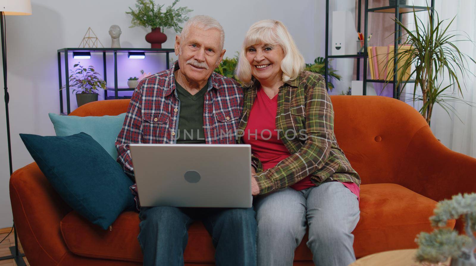 Senior elderly family couple grandparents man woman sitting on couch at home room and opens laptop. Mature grandmother grandfather starts working on computer, sends messages, makes online purchases