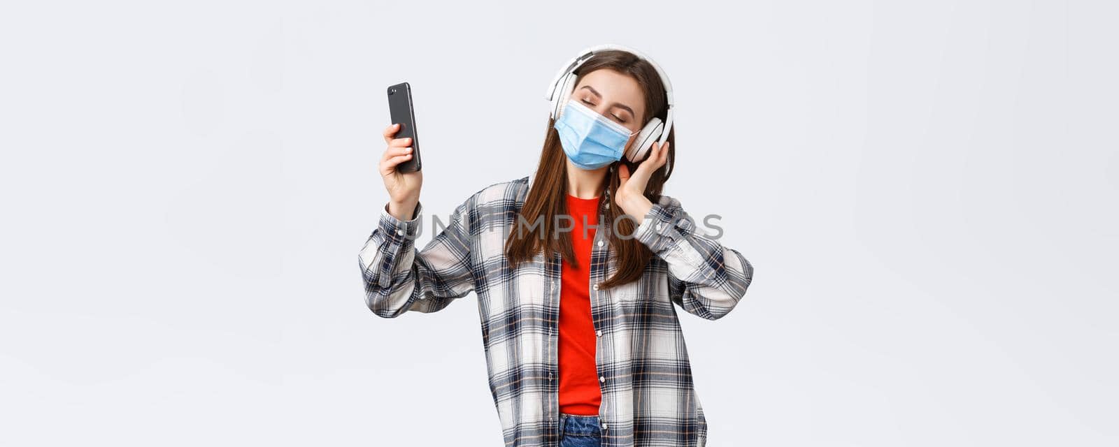 Social distancing, leisure and lifestyle on covid-19 outbreak, coronavirus concept. Carefree young girl in medical mask and headphones, close eyes dancing to music, perfect sound, hold mobile phone by Benzoix