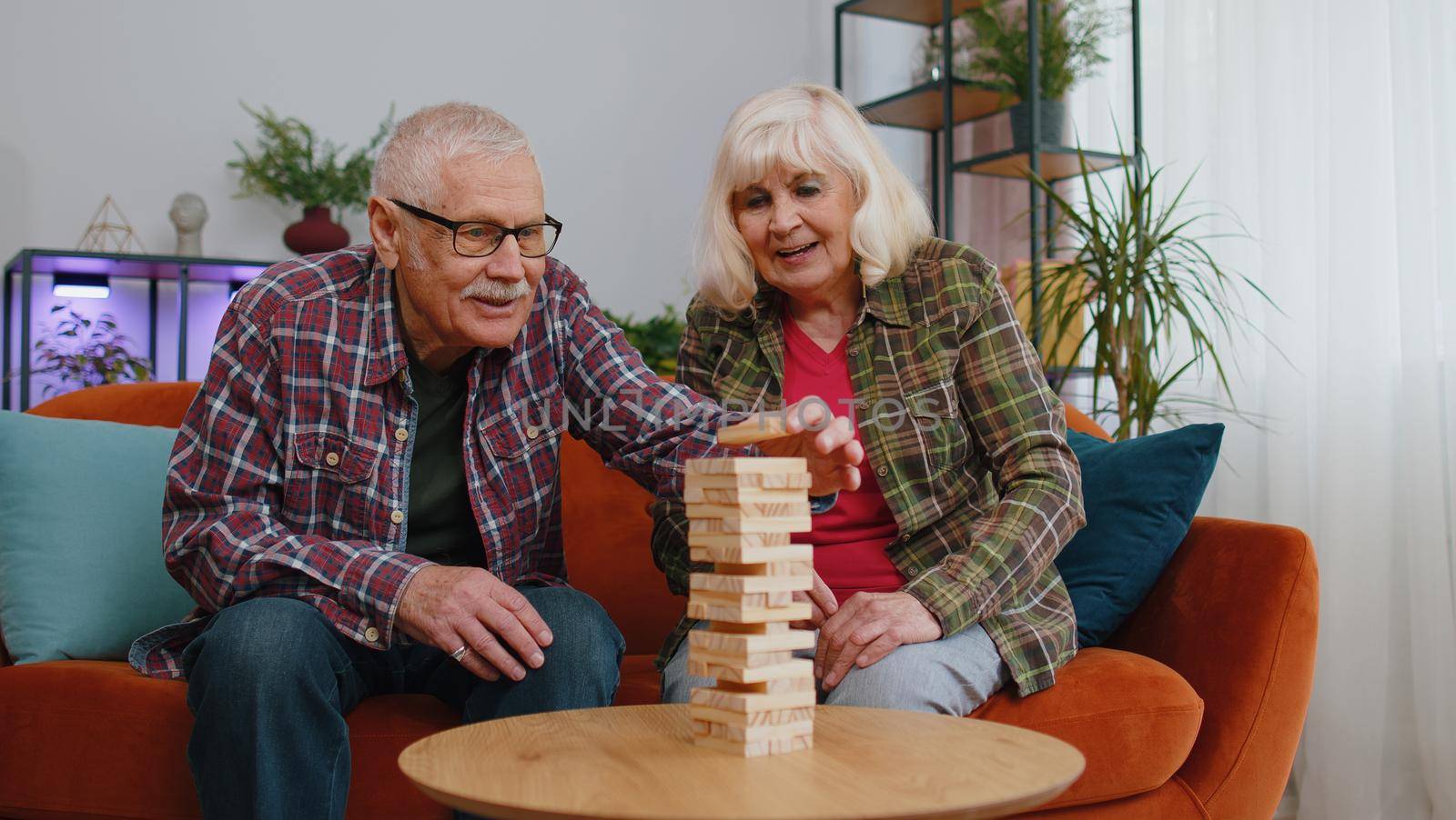 Old senior elderly family couple grandparents man woman playing in blocks build game. Mature grandmother grandfather spending time together play board game hobby with wooden tower on couch at home