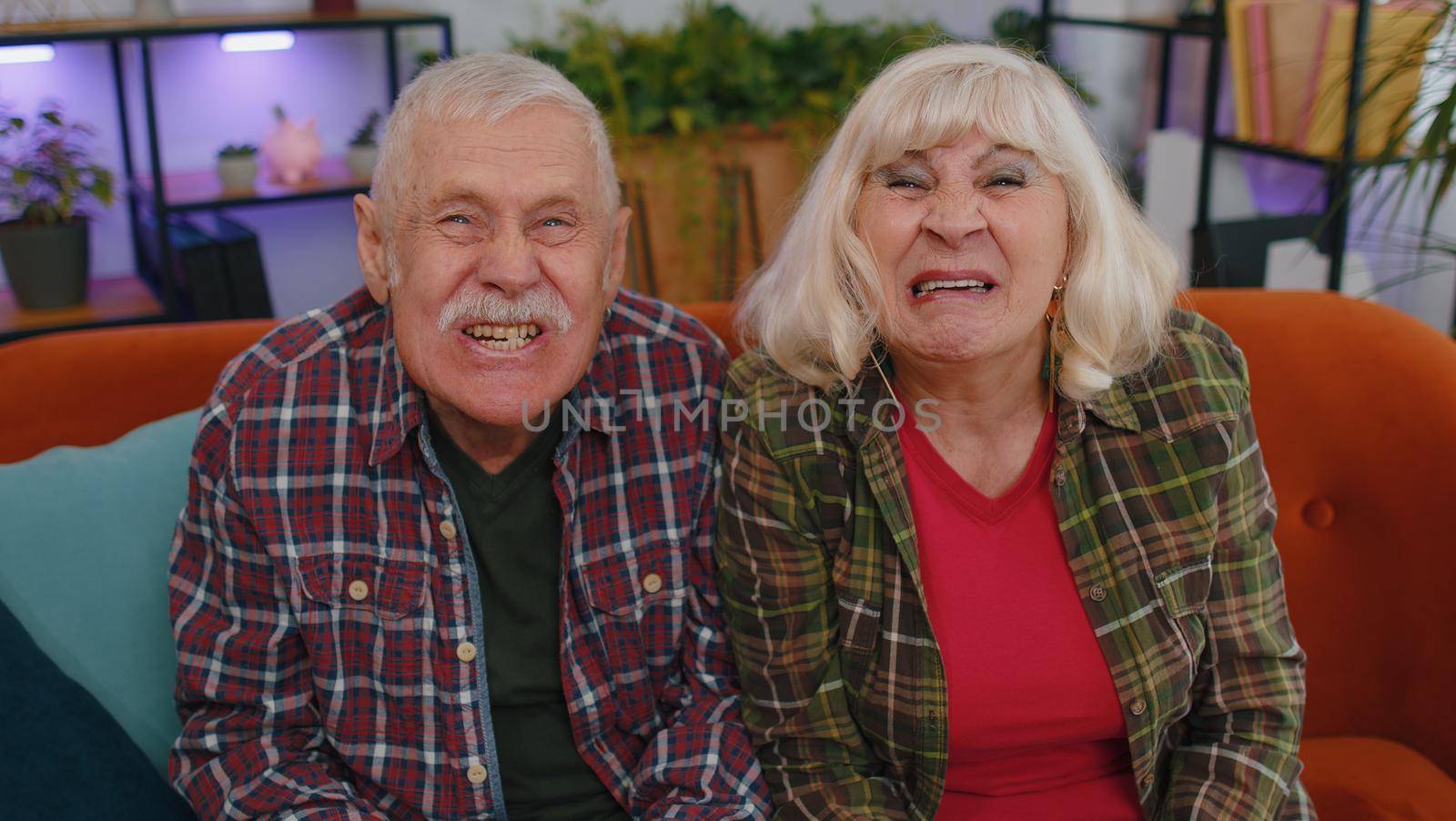Funny old senior elderly family couple grandparents man woman making playful silly facial expressions and grimacing, fooling around, showing tongue. Mature grandmother grandfather on couch at home