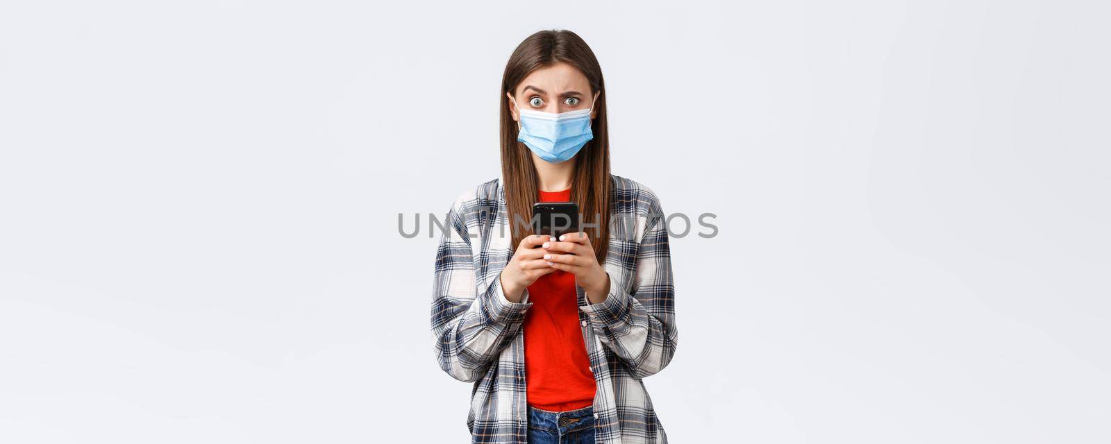 Different emotions, covid-19, social distancing and technology concept. Puzzled and confused young woman in medical mask react to strange message, hold mobile phone, look camera unsure.