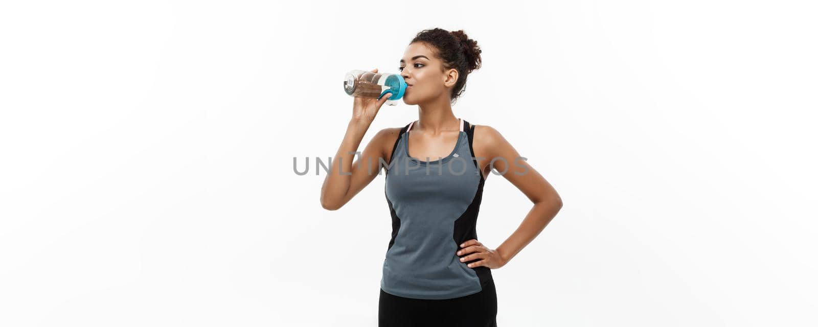 Healthy and Fitness concept - beautiful African American girl in sport clothes drinking water after workout. Isolated on white studio background by Benzoix
