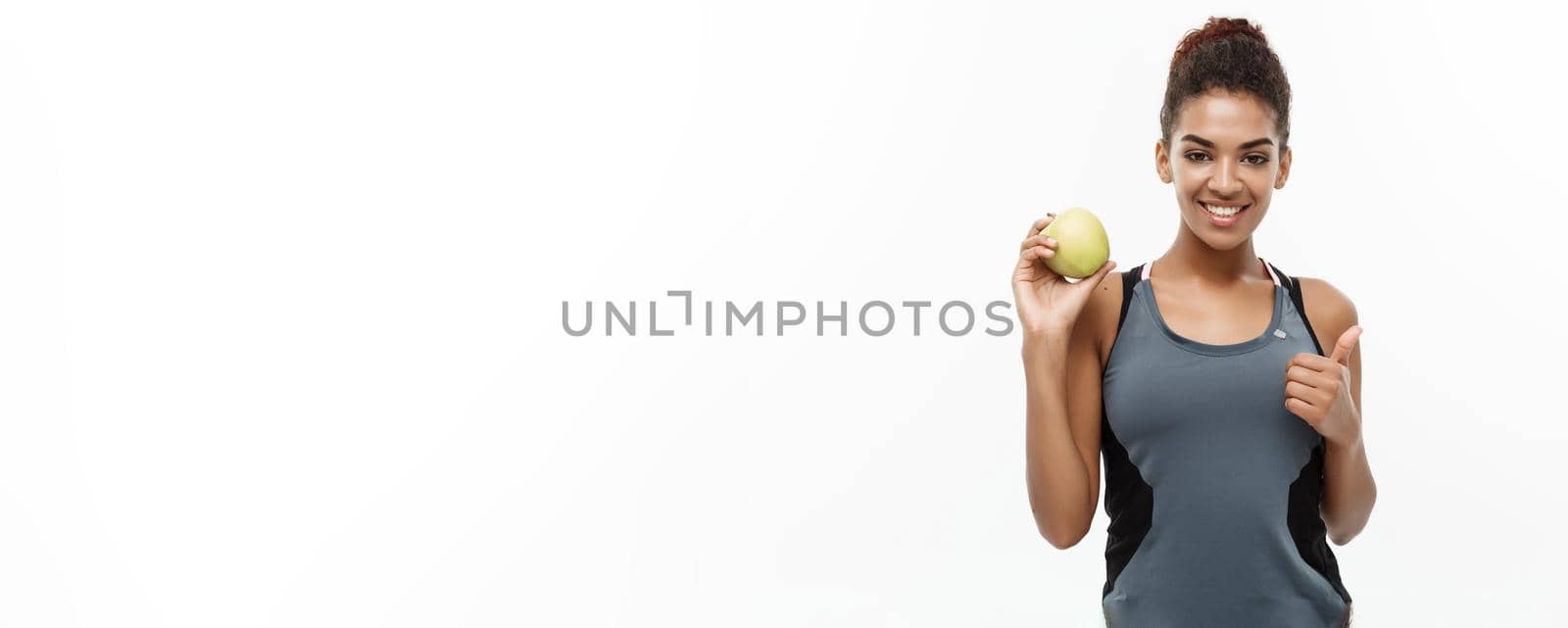 Healthy and Fitness concept - Beautiful American African lady in grey fitness clothes holding green apple. Isolated on white background. by Benzoix