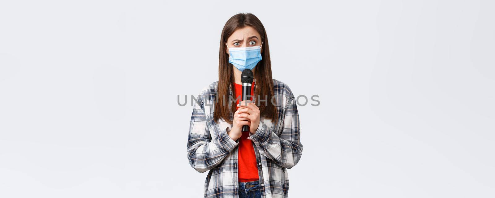 Coronavirus outbreak, leisure on quarantine, social distancing and emotions concept. Confused woman in medical mask look smth strange, holding microphone, perform, white background by Benzoix