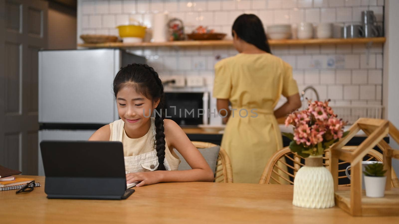 Asian girl having online class on computer tablet, doing homework at kitchen table. Concept of virtual education, homeschooling.