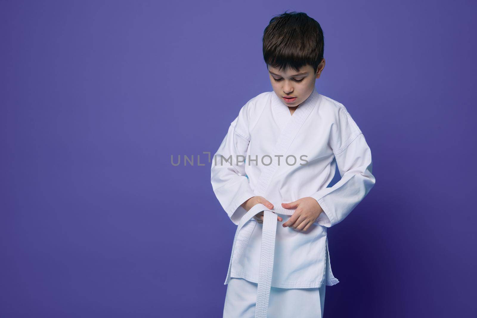 Horizontal portrait of a confident teenage boy aikido fighter tying white belt of his kimono, isolated over violet background with copy ad space. Oriental philosophy and japaneese martial arts concept