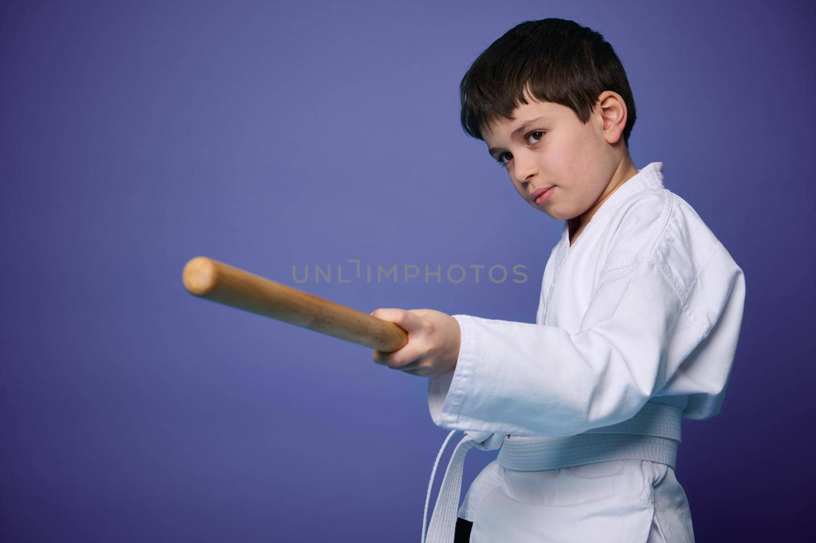 Waist length portrait of a confident teenage boy in white kimono practices wooden bokken in Aikido training on purple background with copy ad space. Concepts of Oriental martial arts practice