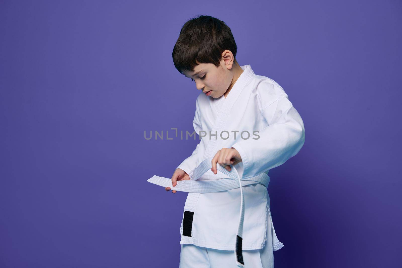 Confident Caucasian teenage boy puts on white kimono, ties a belt around his waist, ready for combat sports. Aikido fighter. Oriental martial arts. Combat sport concept