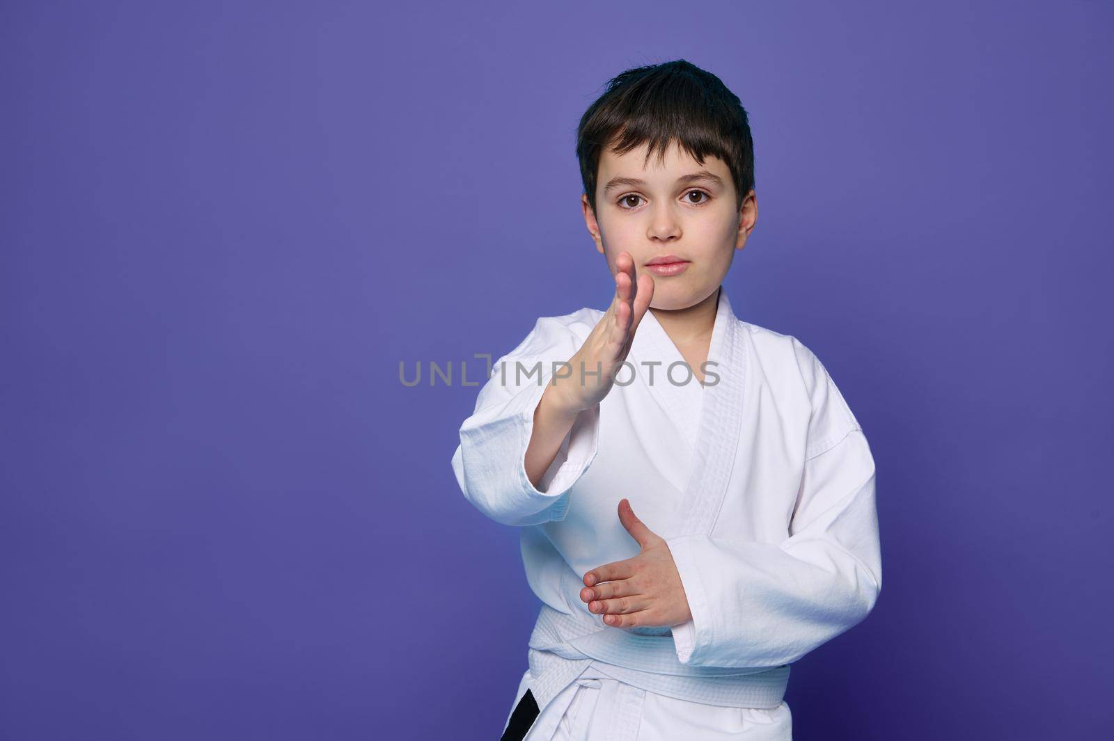 Martial arts attack. Waist up portrait of a school age boy aikido fighter in white kimono isolated over purple background with copy space for advertising text.