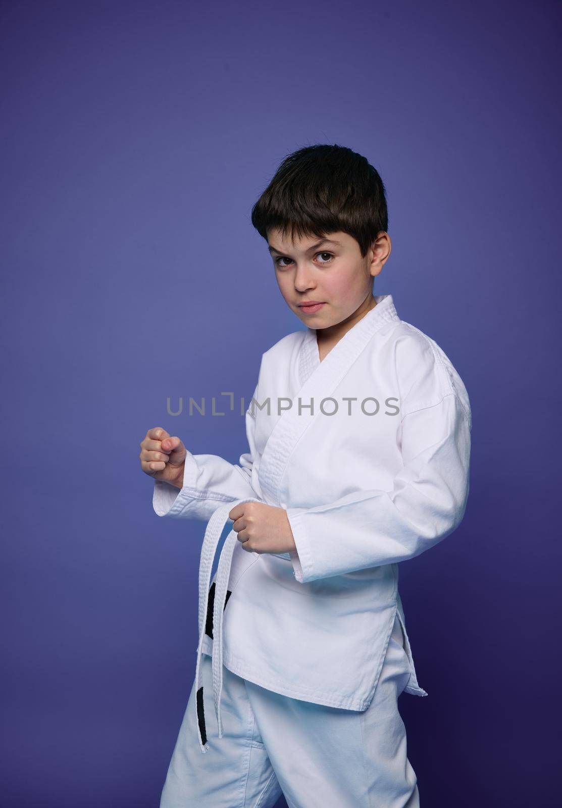 Confident portrait of handsome strong Caucasian teenage boy, aikido fighter practicing martial skills against violet wall background with copy space for advertising text. Oriental martial arts concept