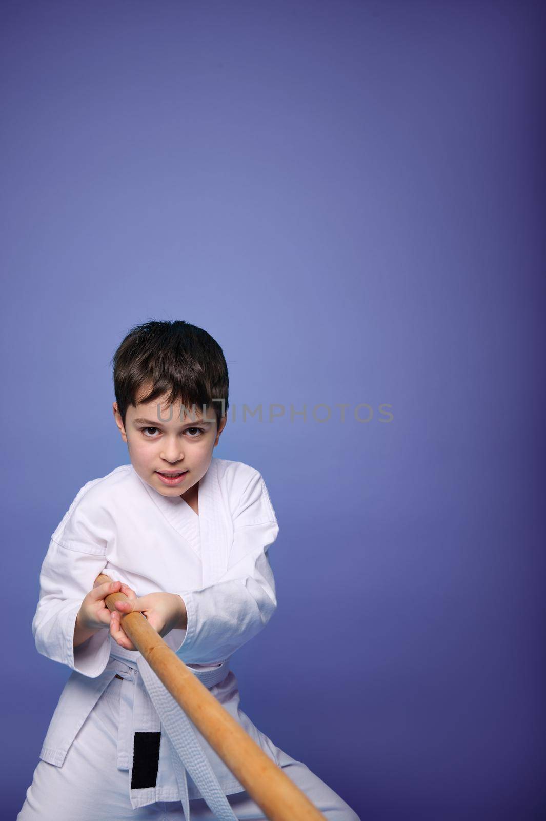 Waist-length portrait of self-confident child boy, teenager, aikido fighter in white kimono, training with wooden weapon - jo bokken sword tanto - practicing oriental martial arts on purple background