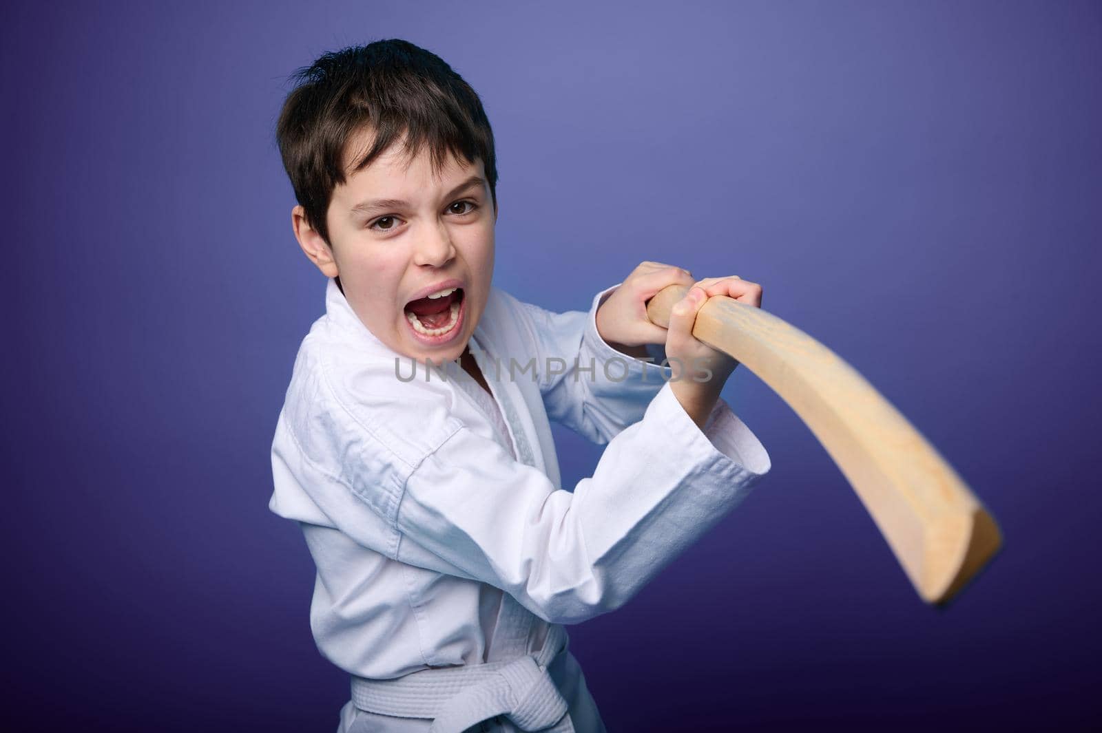 Waist length portrait of a concentrated strong Caucasian teenage boy in white kimono standing in fighting pose with wooden katana sword. Aikido fighter. Oriental martial arts concept