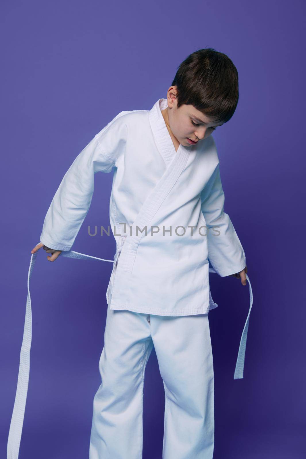 Handsome child, Caucasian teenage boy, aikido wrestler putting on white kimono isolated on purple background with copy space for advertising text. Oriental martial art combat