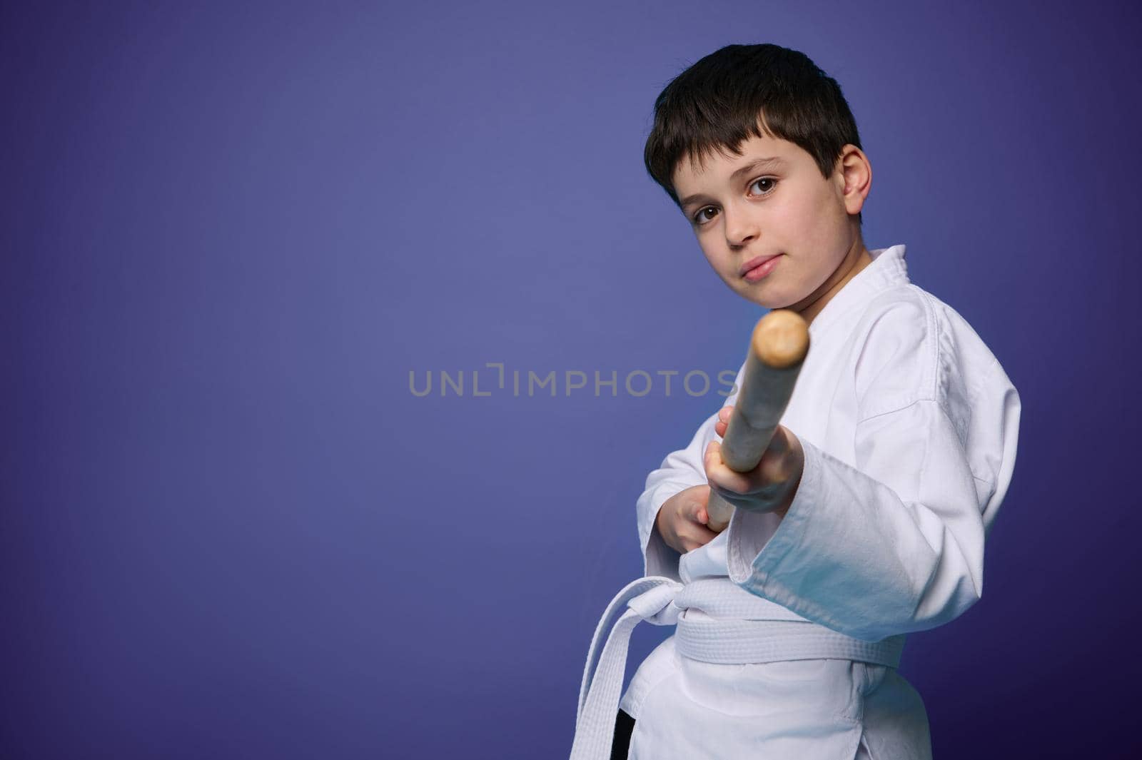 Oriental martial arts practice. Aikido wrestler in white kimono with wooden weapon- bokken practicing fighting skills, looking at camera against purple background with copy space for advertising text
