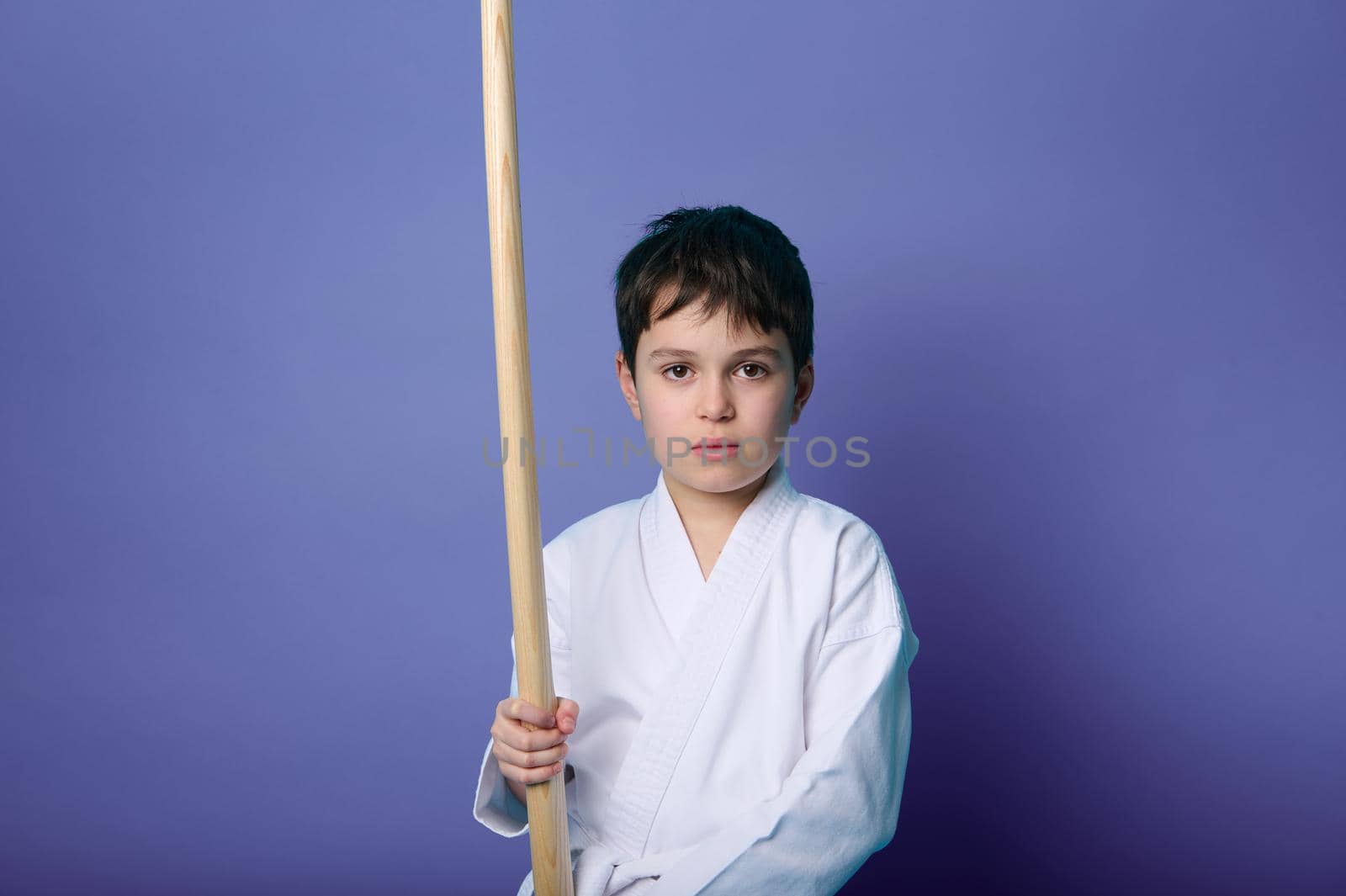 Waist length portrait of charming tranquil confident Caucasian teenage boy martial arts wrestler holding an Aikido wooden sword isolated on purple background with copy space for advertising text