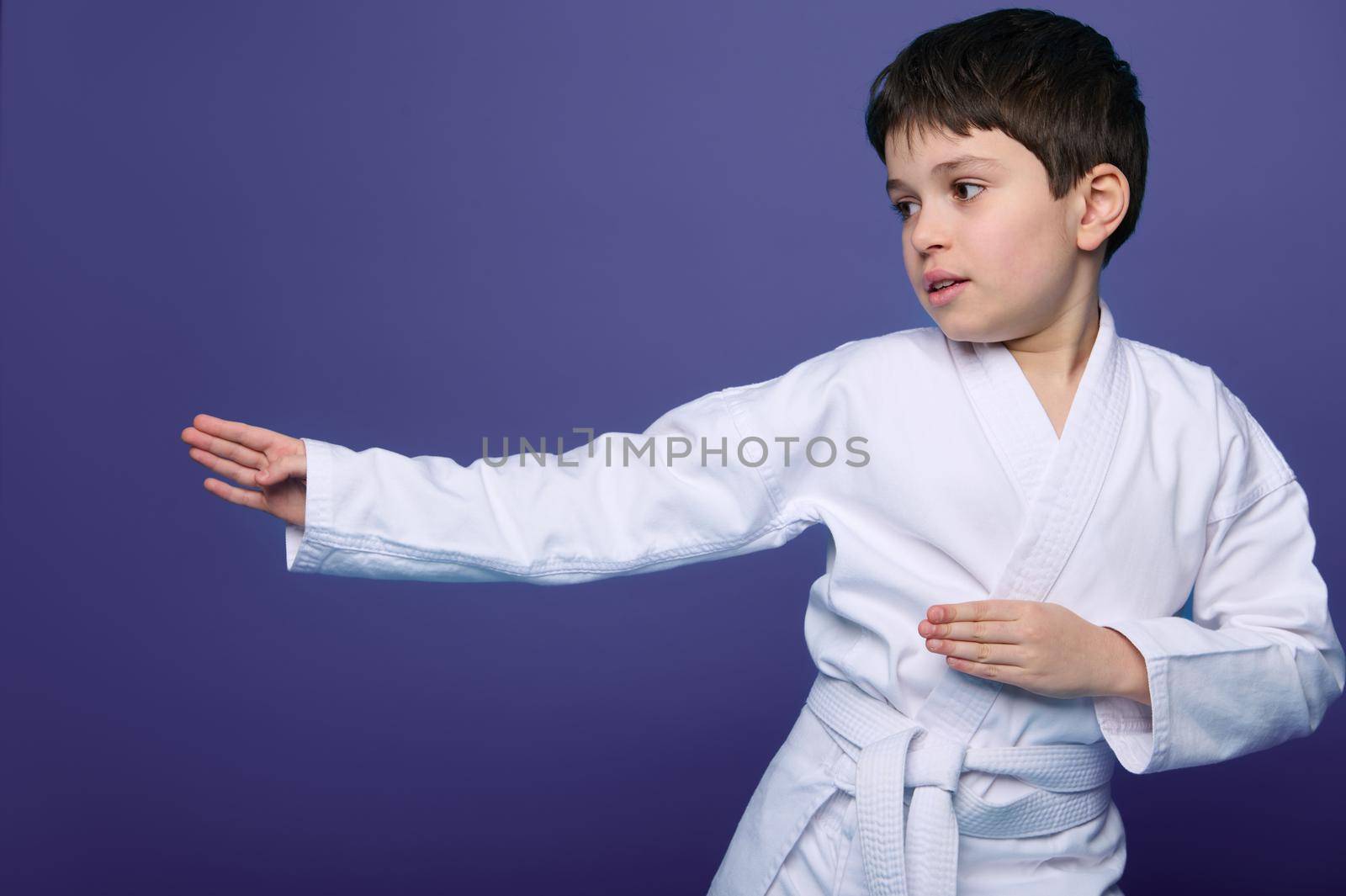 Oriental martial arts. Waist length portrait of an aikido wrestler European 10 years old boy in white kimono improves his fighting skills, isolated on purple background with copy ad space