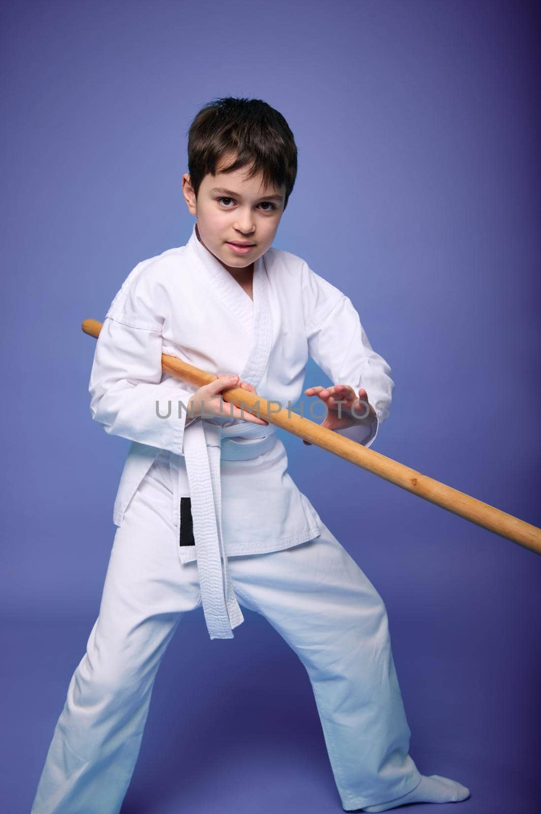 Caucasian teenage boy in a white kimono fights with a wooden sword in aikido training on a purple background with copy space for advertising text. Healthy lifestyle and martial arts concept