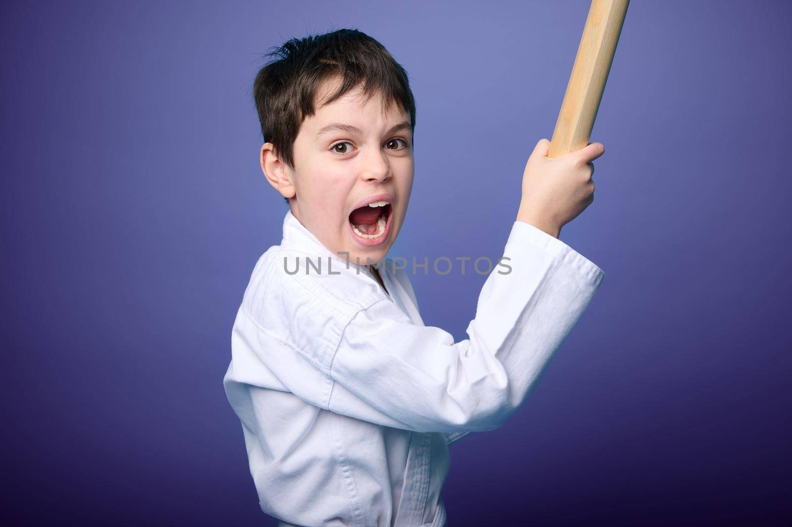 Concentrated strong boy in white samurai hakama kimono - aikido fighter holding bamboo katana weapon and practicing martial skills isolated on purple background with copy space for advertising text