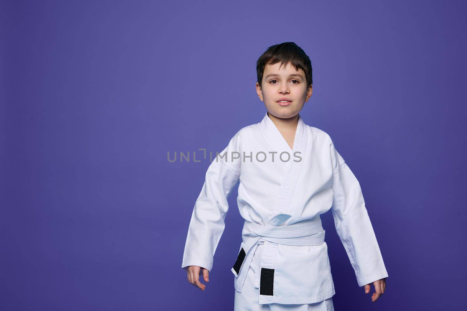 Portrait of charming confident aikido wrestler - Caucasian teenager in white kimono isolated on purple background with copy space for advertising text. Oriental martial arts concept