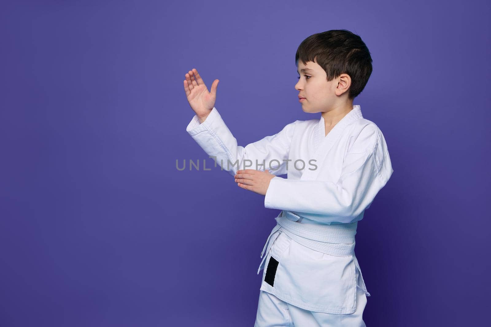 Charming confident Oriental martial arts wrestler - Caucasian school age boy in white kimono- practicing martial skills isolated on purple background with copy space for advertising text. Aikido