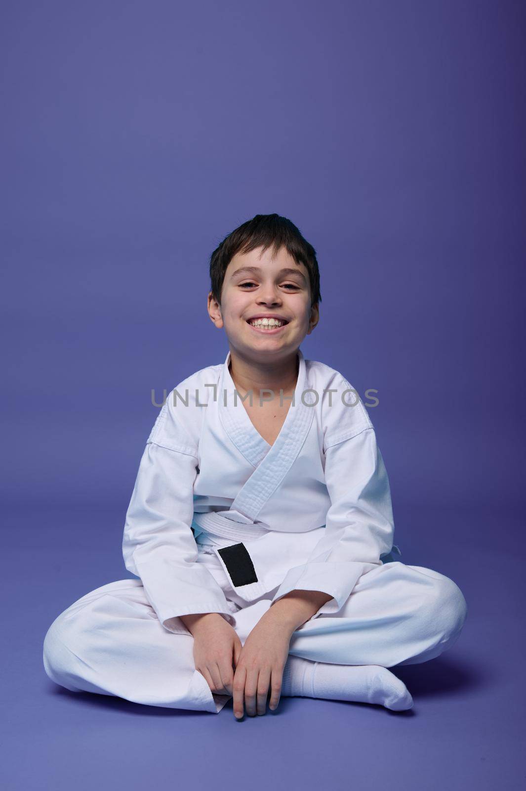 Charming Caucasian teenager - aikido fighter - in white kimono smiles toothy smile, sitting in lotus position while practicing oriental martial arts on purple background with copy ad space for text