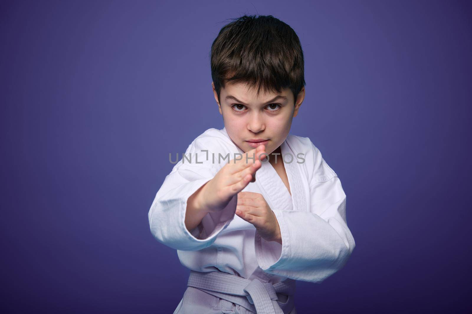 Waist length portrait of a confident strong dark-haired Caucasian kid, aikido fighter practicing martial skills on purple background with advertising copy space. Oriental martial arts practice concept