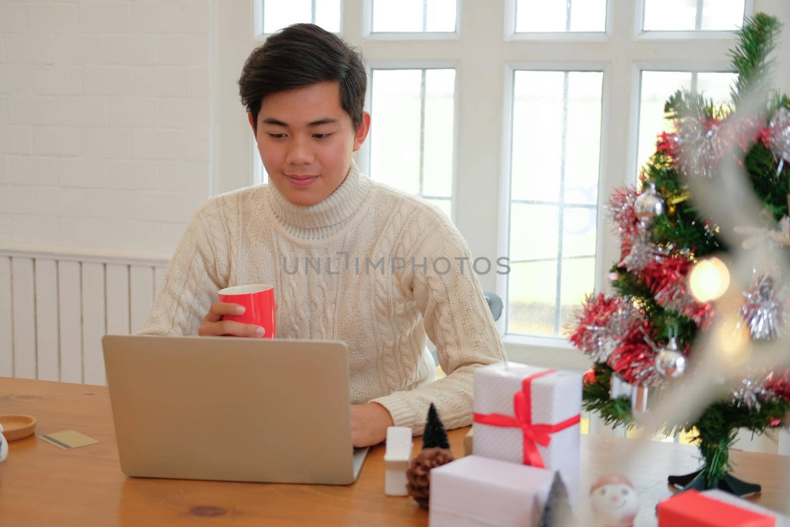 man wearing cream sweater holding coffee cup & using computer at home during christmas. xmas new year celebration