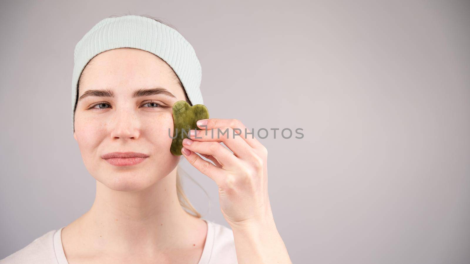 Portrait of a young woman massages her face with a gouache scraper on a white background