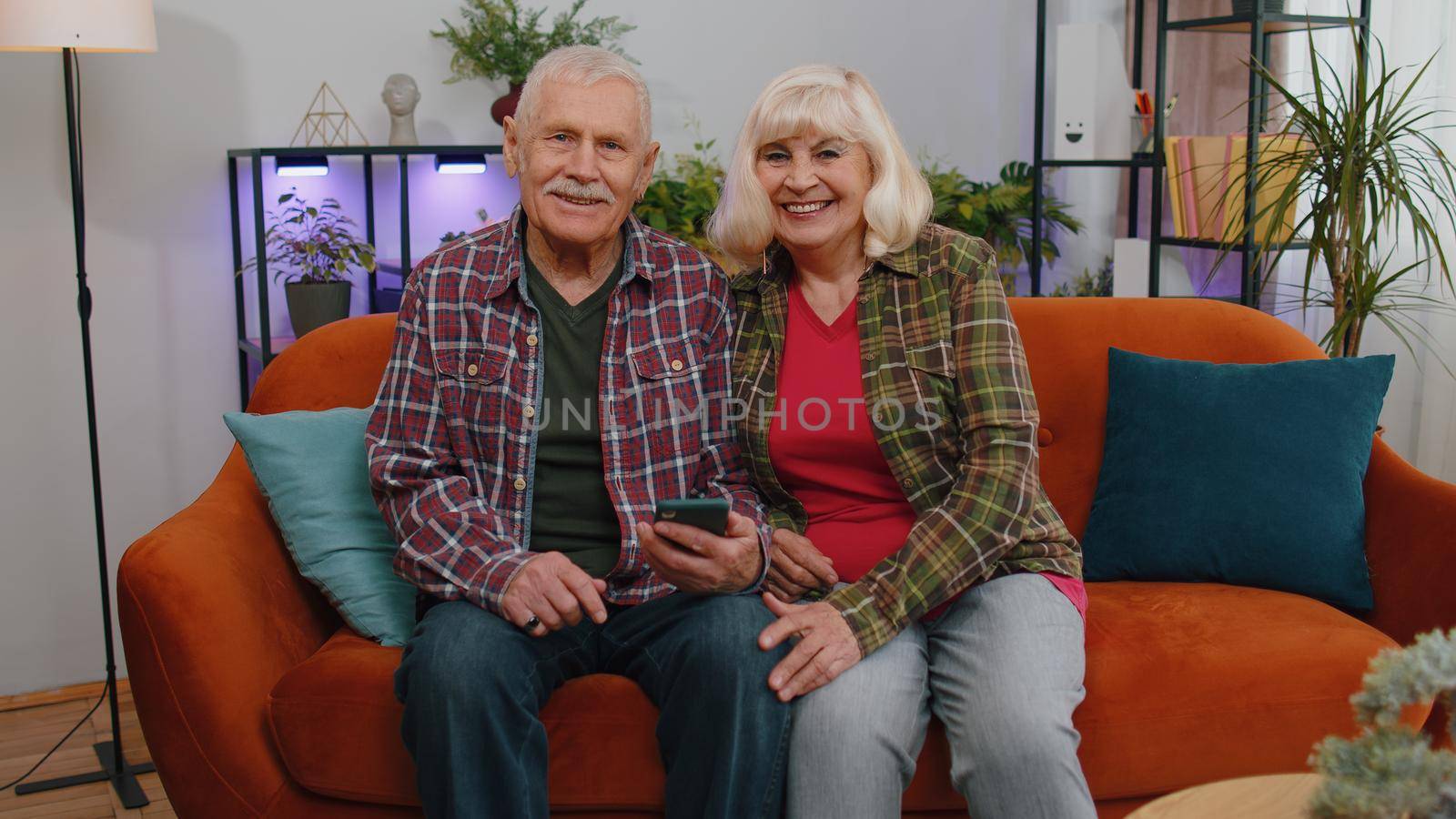 Senior elderly family couple grandparents man woman in home living room and sitting on couch. Mature grandmother grandfather works on smartphone, sends messages, makes online purchases
