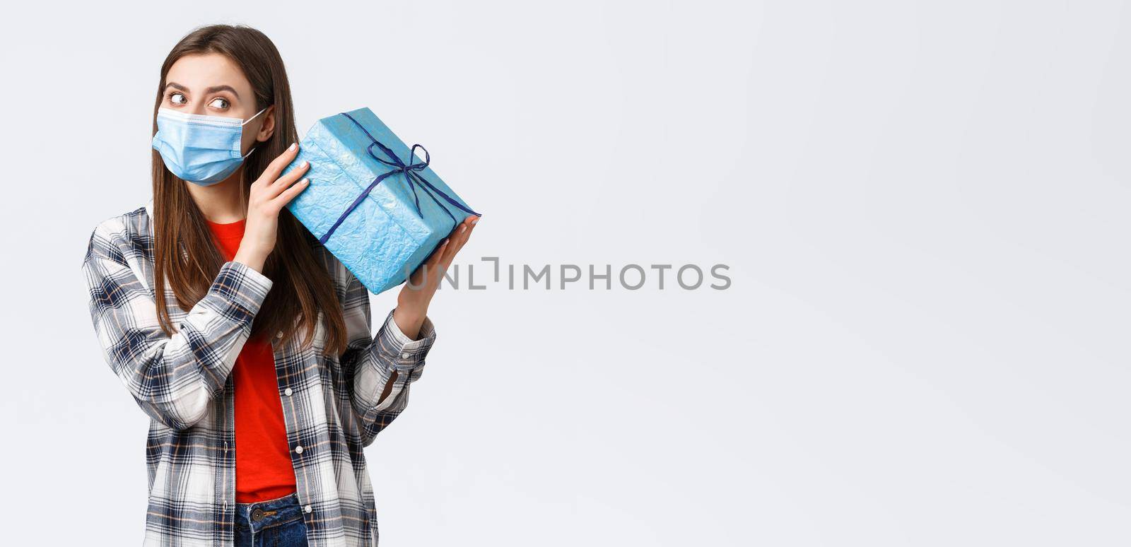 Covid-19, lifestyle, holidays and celebration concept. Cute birthday girl in medical mask, shaking gift box to guess what inside, receive b-day present, celebrating during coronavirus self-quarantine.