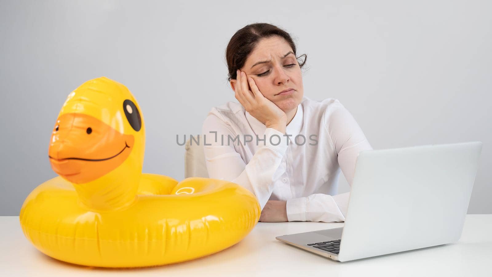 Caucasian woman sits at a table with a laptop and an inflatable duck on a white background. Office worker dreaming of vacation. by mrwed54