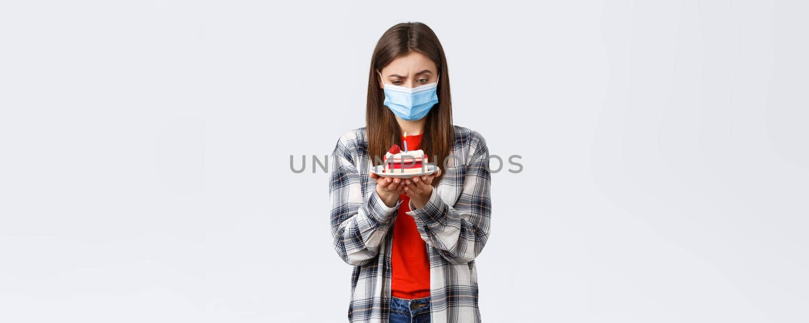 Coronavirus outbreak, lifestyle during social distancing and holidays celebration concept. Confused woman in medical mask staring puzzled at birthday cake lit candle, thinking, white background.