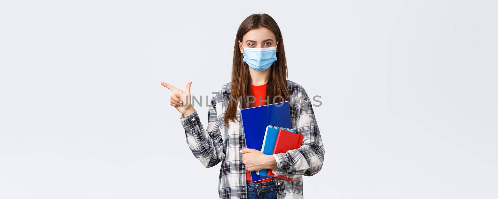 Coronavirus pandemic, covid-19 education, and back to school concept. Young pretty female student in medical mask with notebooks, pointing finger left, showing university info by Benzoix