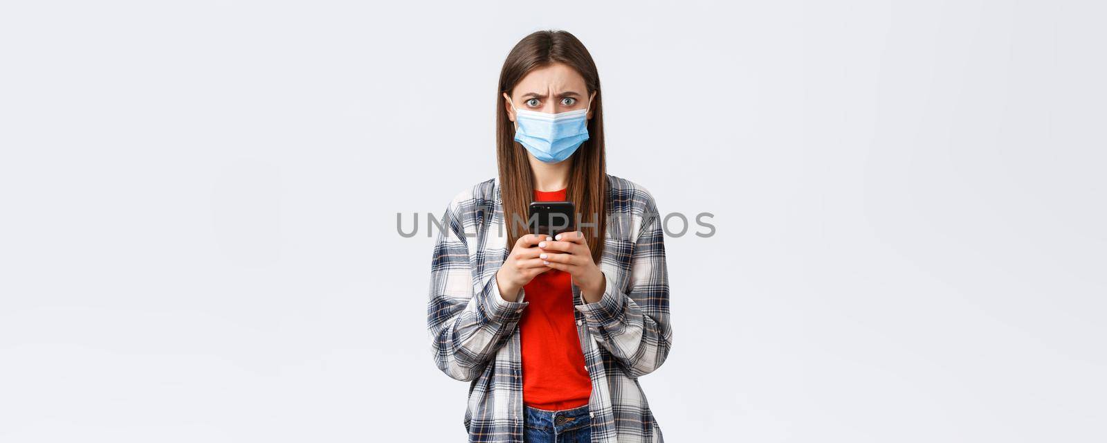 Different emotions, covid-19, social distancing and technology concept. Frustated and confused young woman in medical mask react to strange message, hold mobile phone, frowning camera.