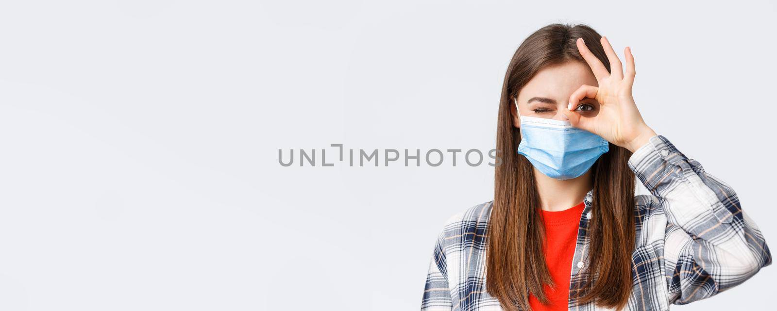 Coronavirus outbreak, leisure on quarantine, social distancing and emotions concept. Close-up of optimistic good-looking woman in medical mask show okay sign and wink.