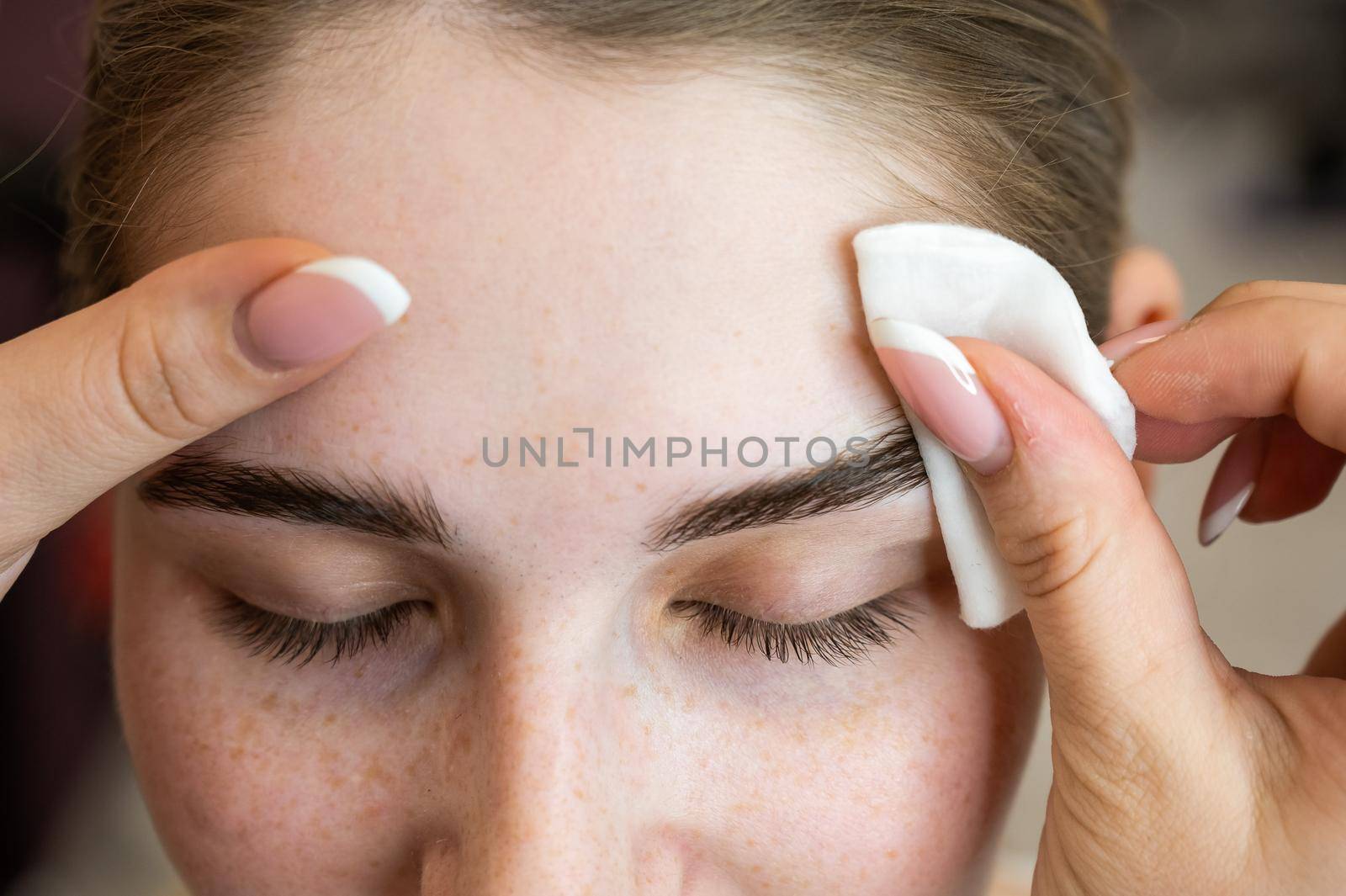 The master processes the eyebrows of a Caucasian woman with a cotton pad before laminating the eyebrows