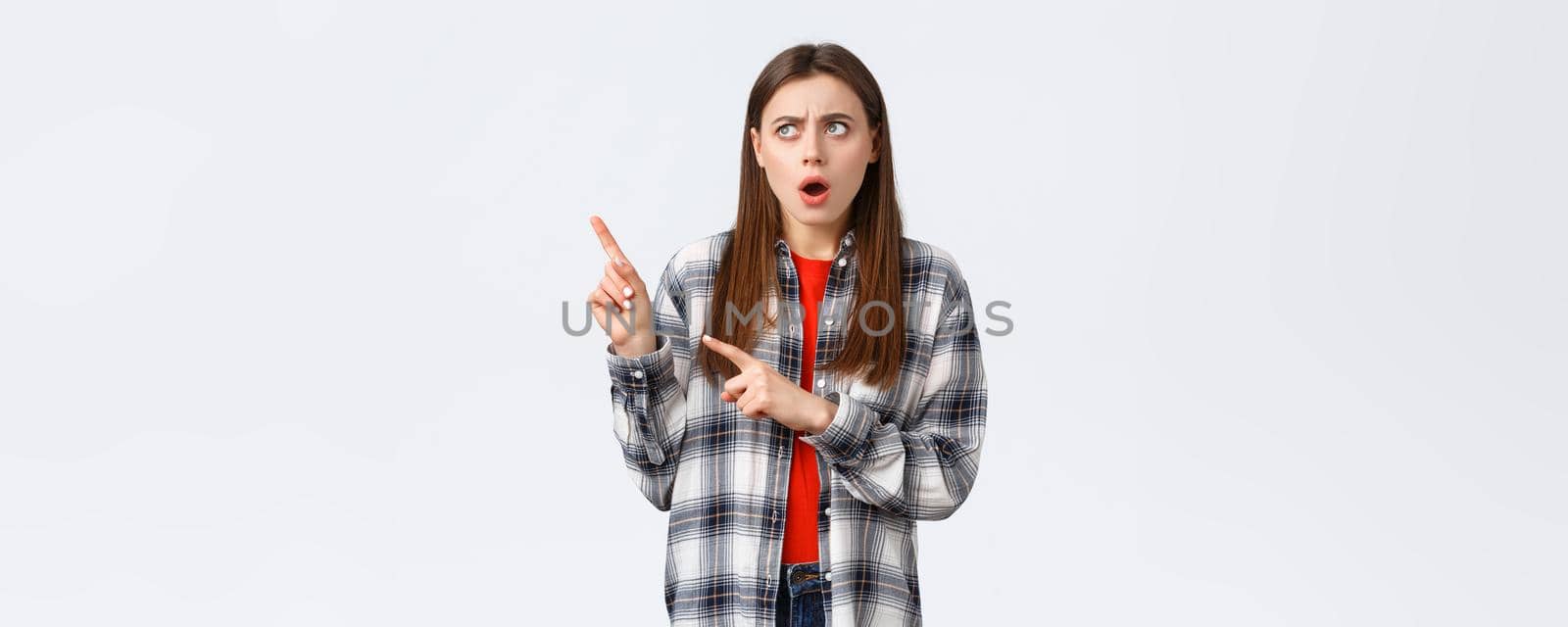 Lifestyle, different emotions, leisure activities concept. Shocked and concerned young pretty woman in checked casual shirt, pointing and looking upper left corner with displeased shook face.