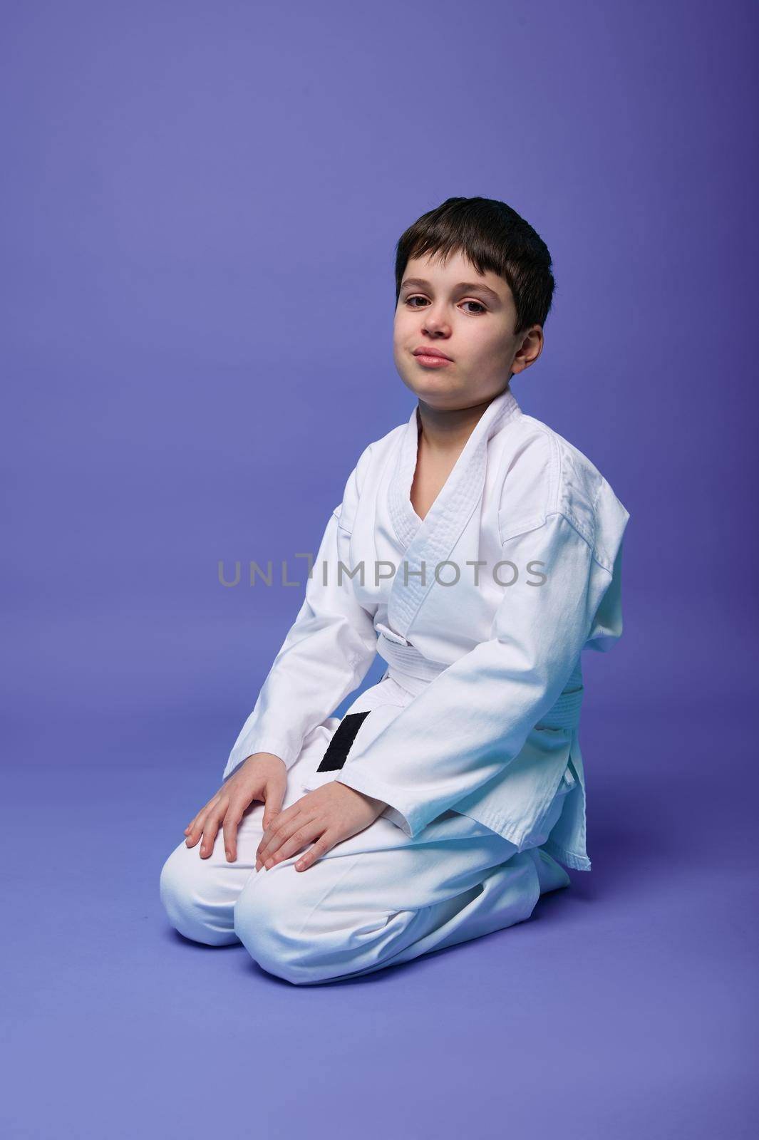 Isolated portrait on violet background of a handsome tranquil confident Caucasian teenage boy - aikido fighter in a white kimono during the practice of oriental martial arts.