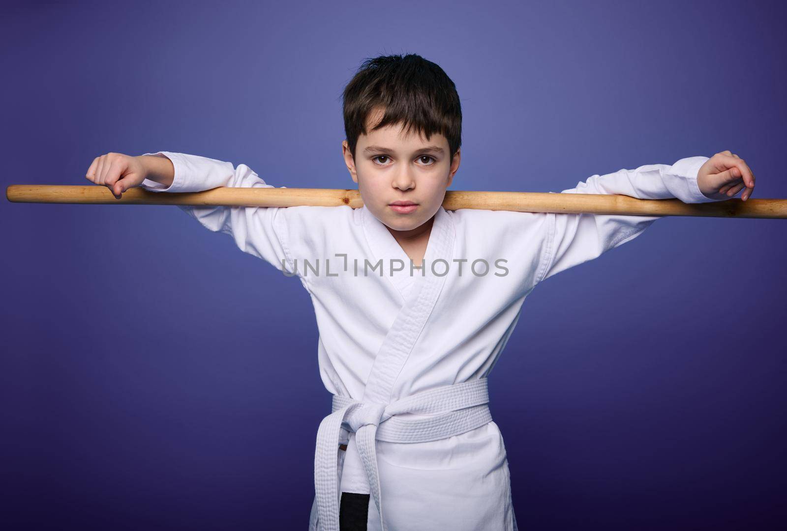 Confident charming European aikido wrestler posing with wooden weapon against violet background with copy ad space for advertising text. Oriental martial arts practice