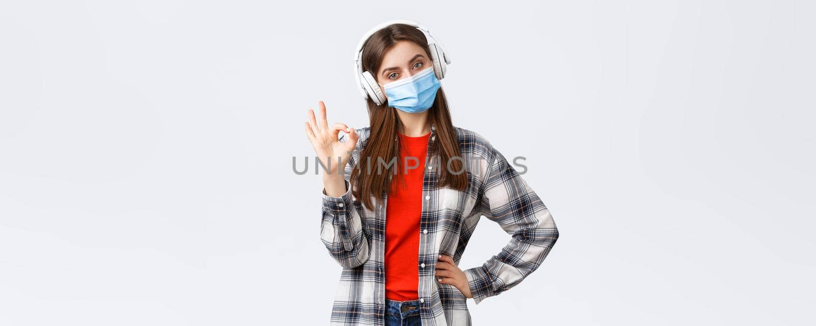 Social distancing, leisure and lifestyle on covid-19 outbreak, coronavirus concept. Pleased good-looking woman in medical mask and headphones, listening music, show okay sign, approve or like.