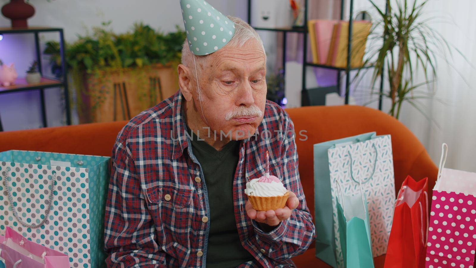 Happy old senior elderly man wears festive birthday hat holds cupcake makes wish joyful congratulating blowing burning candle on cake. Mature grandfather celebrating party on couch with gifts at home