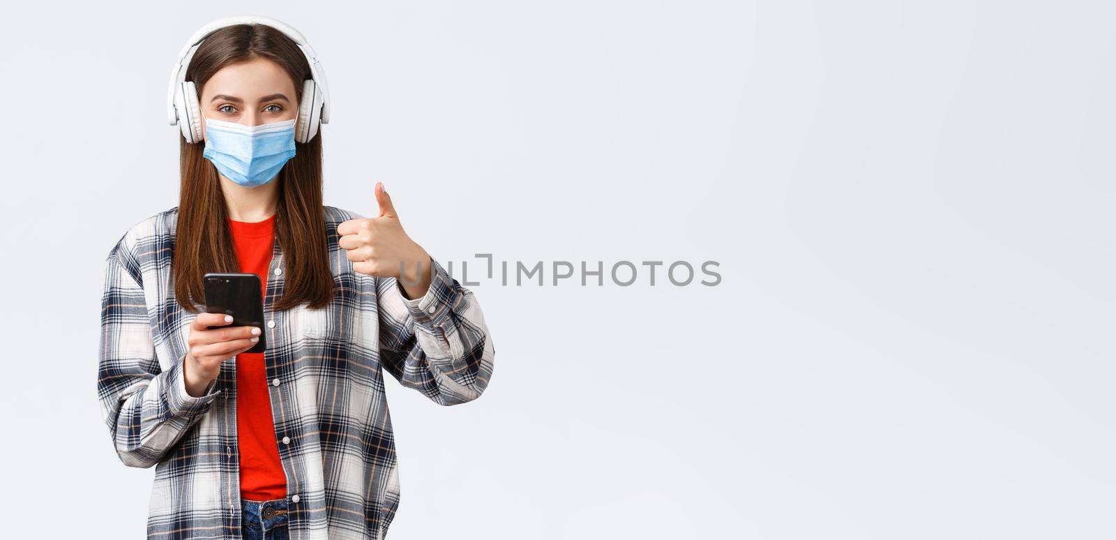 Social distancing, lifestyle on covid-19 outbreak, coronavirus concept. Cheerful pleased good-looking woman in medical mask, approve nice sound of wireless headphones, thumb-up, hold phone.